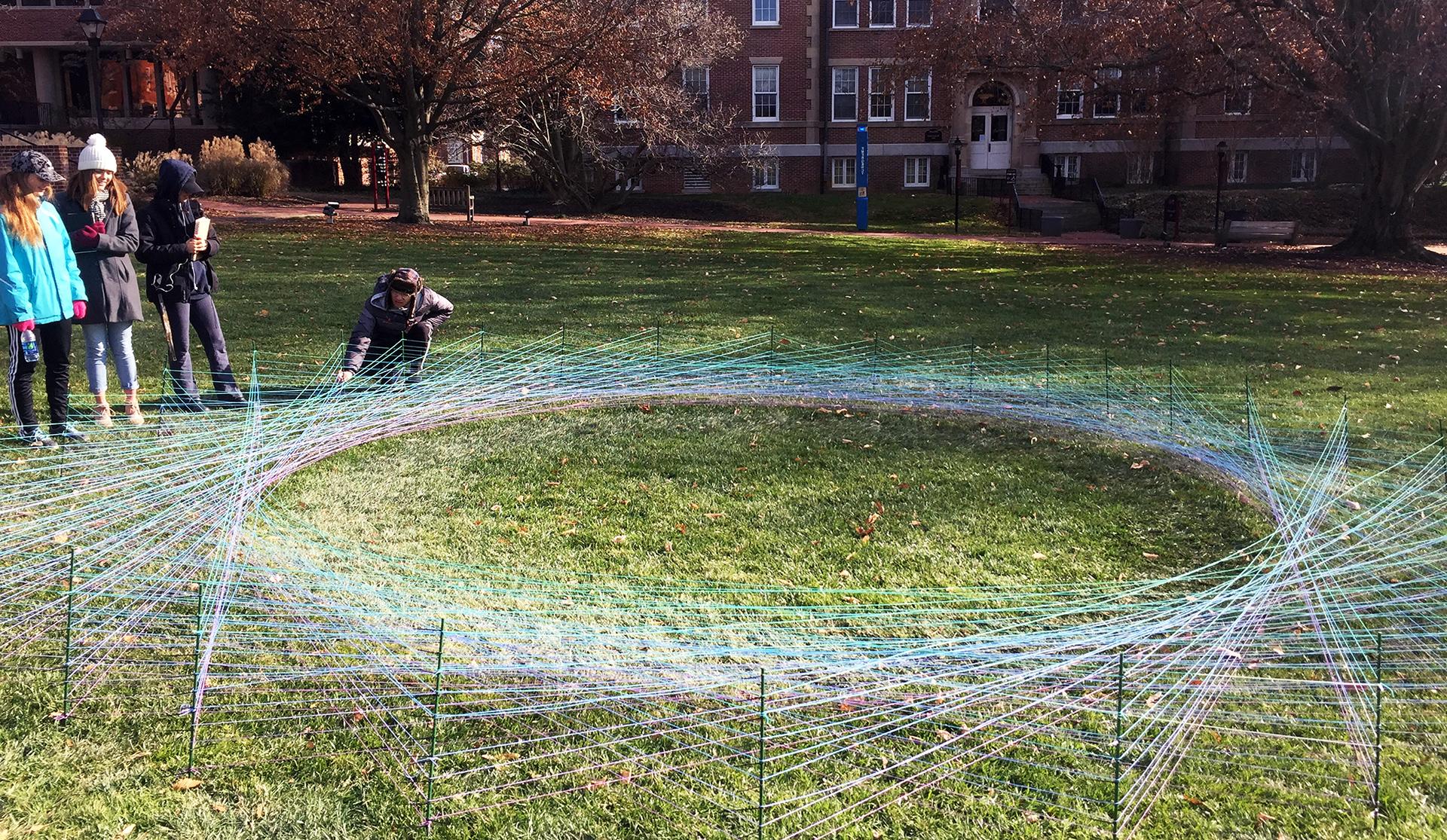 Students gathered around a student artwork made of colorful intersecting strings, in front of Bunting Hall on the WC campus